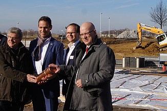 Vier Männer auf der Baustelle in Schönfeld mit symbolischer Grundsteinlegung. Sie halten ein Gefäß.