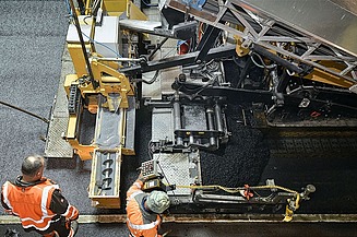 Foto zeigt zwei Straßenbauarbeiter in Schutzwesten vor einer Asphaltbaumaschine