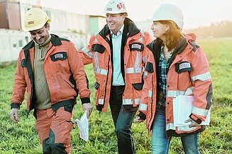 Foto von drei Personen in Arbeitskleidung auf einer Baustelle, die gut gelaunt wirken und Unterlagen bei sich tragen