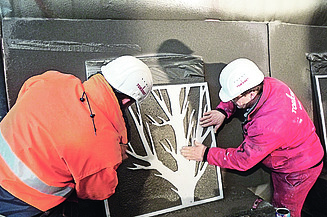  Foto von zwei Männern die eine Schablone von einem Baum auf eine braune Platte halten.