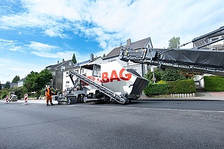 Foto einer Asphaltbaumaschine mit der Aufschrift STRABAG, sie steht am Straßenrand, der Himmel ist bewölkt