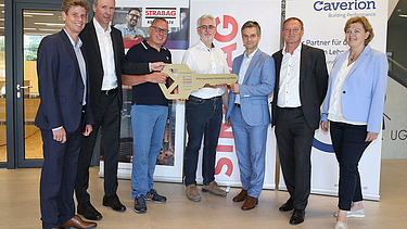 Foto von sieben Personen bei der symbolischen Schlüsselübergabe, sie stehen vor zwei Roll-ups