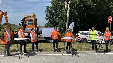 Foto von mehreren Personen in Schutzwesten bei einer Baustellenbesichtigung am Straßenrand stehend