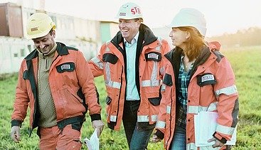 Foto von drei Personen in Arbeitskleidung auf einer Baustelle, die gut gelaunt wirken und Unterlagen bei sich tragen