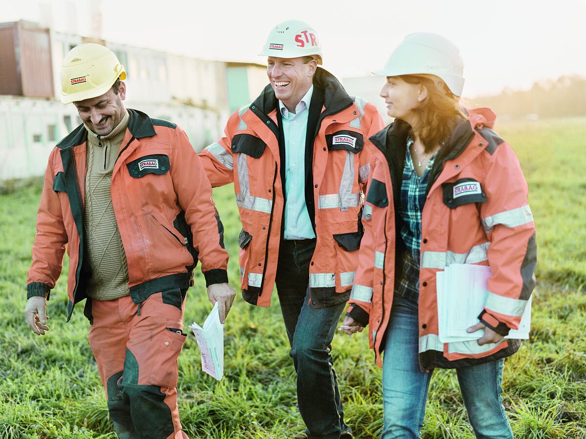 Foto von drei Personen in Arbeitskleidung auf einer Baustelle, die gut gelaunt wirken und Unterlagen bei sich tragen