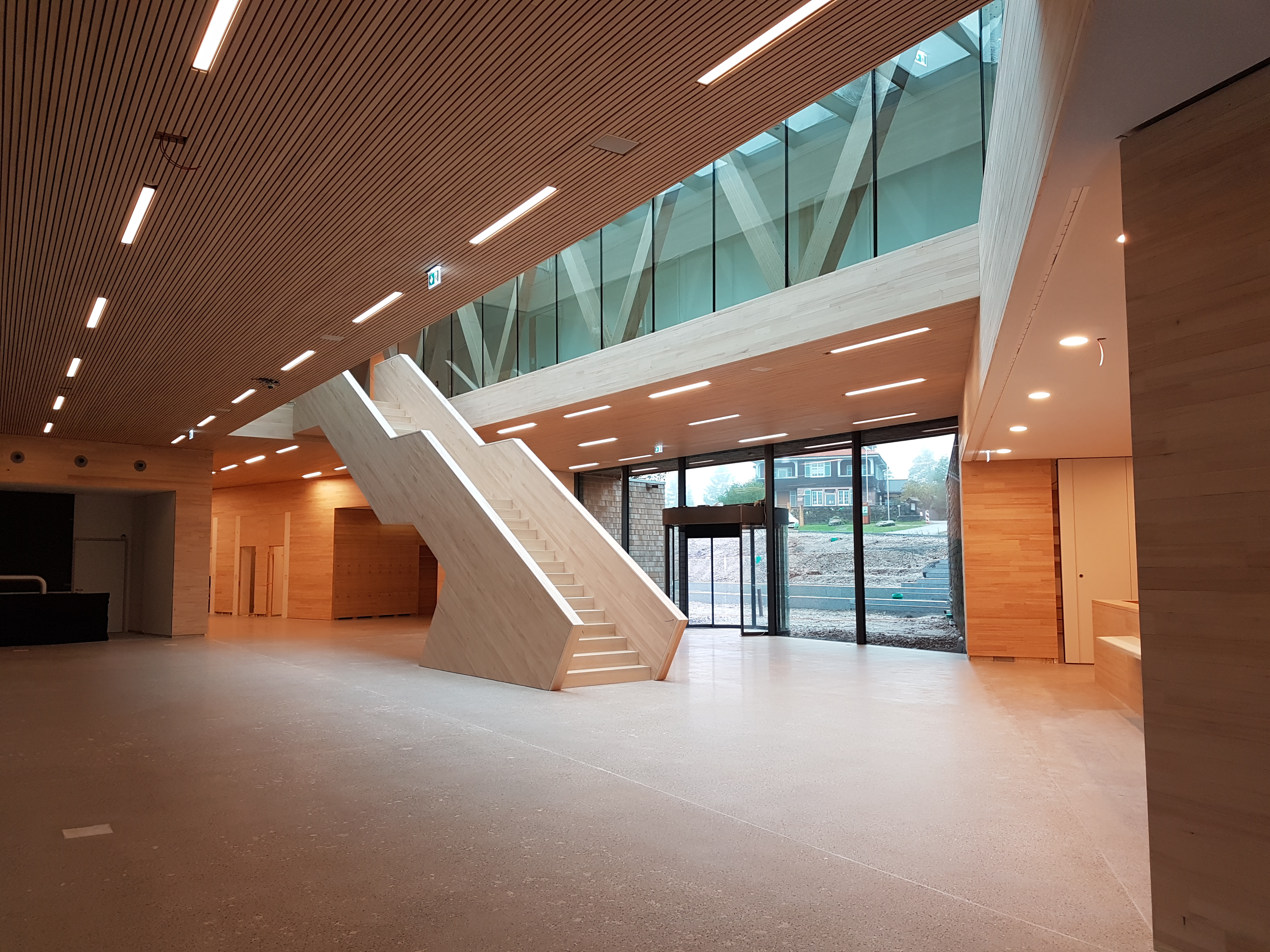 Foto vom inneren eines Gebäudes in der Eingangshalle. In der Mitte erstreckt sich eine Treppe aus Holz.  