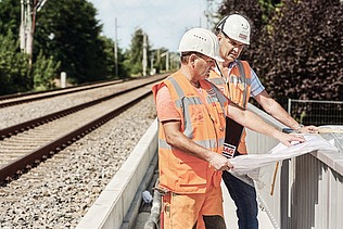 Foto von einem Bahngleis, rechts stehen zwei Personen in Schutzkleidung, die einen Plan in der Hand halten