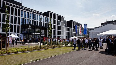 Foto vom Unternehmenszentrale in Köln auf dem Parkplatz. Es ist ein Zelt aufgebaut und Menschen feiern die neue Zentrale.   