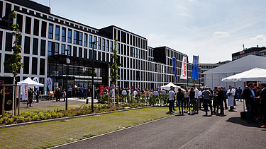 Foto vom Unternehmenszentrale in Köln auf dem Parkplatz. Es ist ein Zelt aufgebaut und Menschen feiern die neue Zentrale.   
