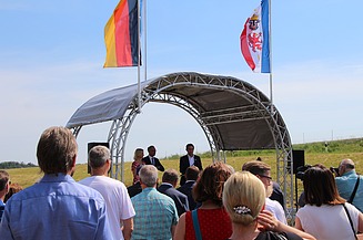 Foto von mehreren Personen, die vor einer überdachten Bühne stehen, dahinter zwei Flaggen