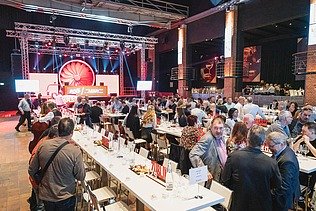 Foto von einer großen Anzahl an feiernden Personen in einem Festsaal, sitzend an weißgedeckten Tischen, im Hintergrund eine Bühne mit Scheinwerfern