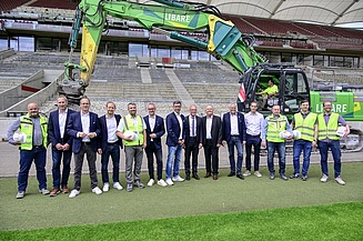 Foto von mehren Personen am Rand eines Fußballplatzes. Hinter ihnen steht ein Bagger. 