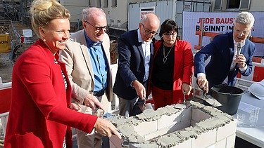Foto von mehreren Personen, die schick gekleideten sind, die Beton auf ein Gemäuer verstreichen. 