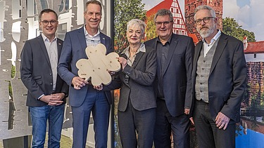 Photo of five people standing in front of a picture wall, the second from the left holding a large wooden clover leaf in her hand