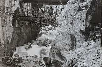 Schwarz-weiß Foto von einer Brücke, die über einen Graben geht. Drumherum sind Bergwände, die sich zu einer Schlucht formen. Auf der Brücke sind zwei Personen zu sehen. 