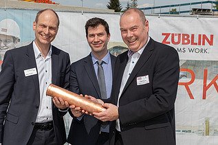 Drei Männer schauen freundlich und halten eine Röhre. 