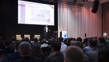 Foto von einem verdunkeltem Veranstaltungsraum, in dem mehrere Personen sitzen, im Hintergrund eine große Projektion