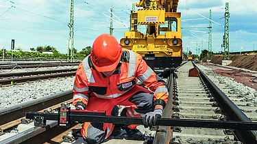 Foto eines Schienenarbeiters in Schutzkleidung, dahinter ein gelbes Schienenbaufahrzeug