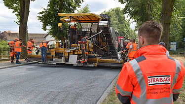 Foto einer Baustelle mit einer Aspahltbaumaschine, im Vordergrund steht eine Person in einer orangen Schutzweste