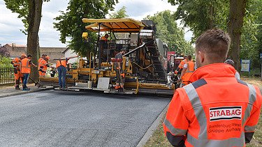 Foto einer Baustelle mit einer Aspahltbaumaschine, im Vordergrund steht eine Person in einer orangen Schutzweste