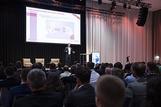 Foto von einem verdunkeltem Veranstaltungsraum, in dem mehrere Personen sitzen, im Hintergrund eine große Projektion