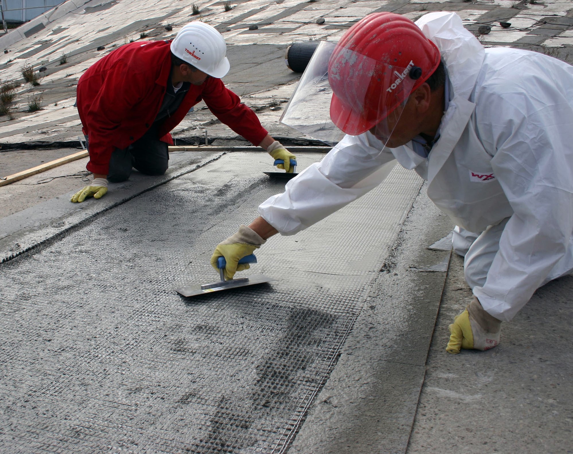  Foto von zwei Personen die mit Spachteln ein Gitter mit Beton bestreichen.
