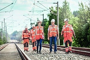 Foto von fünf Personen, die zwischen zwei Bahngleisen mit Oberleitungen gehen