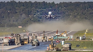 Foto einer Baustelle auf einer Flugpiste, dahinter startet gerade ein Flugzeug