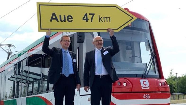 Das Bild zeigt zwei Personen im Anzug, die eine gelbe Verkehrstafel in die Höhe halten, Dahinter steht eine Straßenbahn