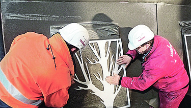  Foto von zwei Männern die eine Schablone von einem Baum auf eine braune Platte halten.