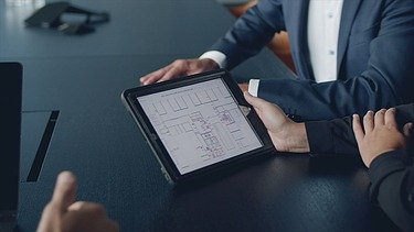 Foto von einem einer Besprechung mit einem Fokus auf ein I-Pad. Auf dem I-Pad ist ein Bauplan zusehen.  