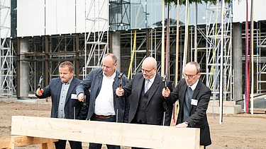Foto von vier Männern die einen Nagel in ein Holzbrett hauen.  