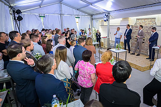 Foto von drei Personen auf einer Bühne bei der Grundsteinlegung des Franzoesischen Saals, davor stehen eine Ansammlung von vielen Personen