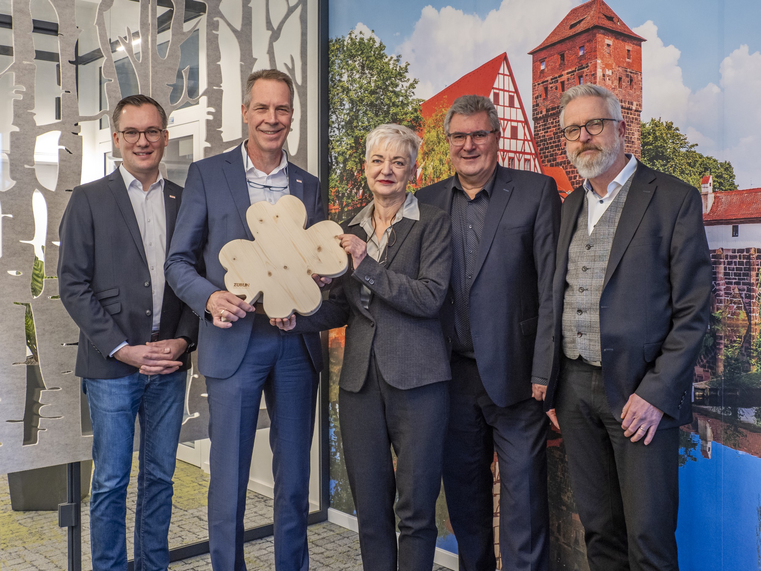 Photo of five people standing in front of a picture wall, the second from the left holding a large wooden clover leaf in her hand