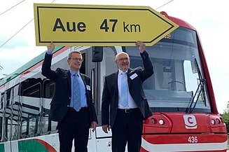 Das Bild zeigt zwei Personen im Anzug, die eine gelbe Verkehrstafel in die Höhe halten, Dahinter steht eine Straßenbahn