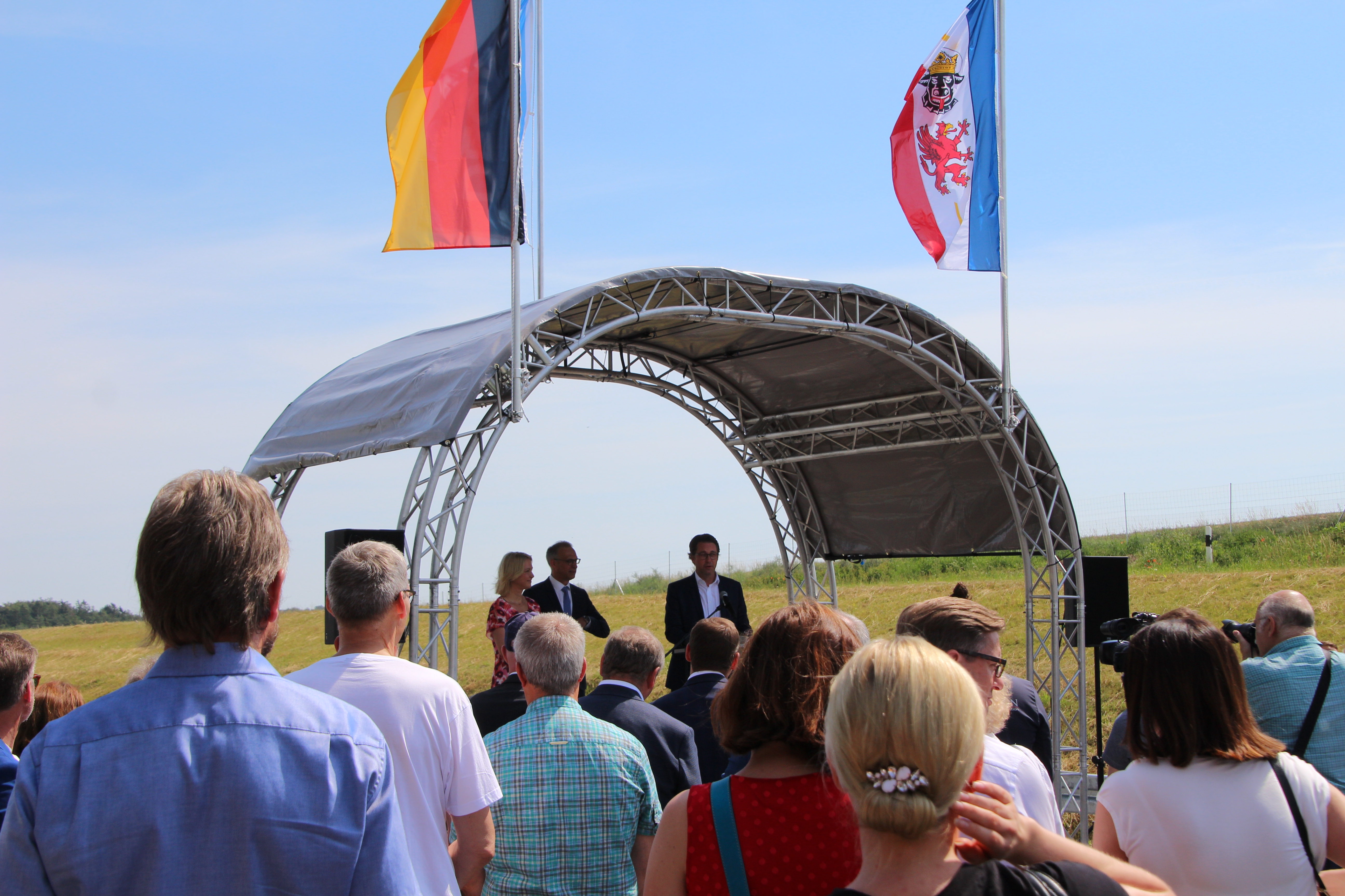 Foto von mehreren Personen, die vor einer überdachten Bühne stehen, dahinter zwei Flaggen