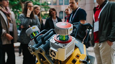 Foto zeigt im Vordergrund einen Motor, dahinter stehen einige Personen, die ihn betrachten