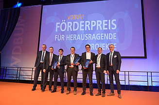 Foto von mehreren Männern in Anzug auf einem Bühnen. Im Hintergrund ein Banner vom VDBUM: „Förderpreis für Herausragende Projekte“ zu sehen.            