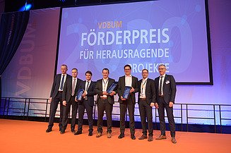 Foto von mehreren Männern in Anzug auf einem Bühnen. Im Hintergrund ein Banner vom VDBUM: „Förderpreis für Herausragende Projekte“ zu sehen.            