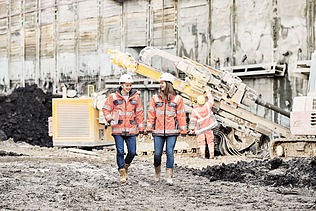Foto von einem Mann und einer Frau. Beide schauen glücklich. Sie befinden sich auf einer Baustelle. Hinter ihnen steht eine große Maschine.   
