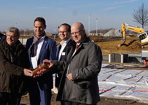 Vier Männer auf der Baustelle in Schönfeld mit symbolischer Grundsteinlegung. Sie halten ein Gefäß.