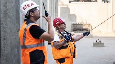 Foto von zwei Arbeitern, der vordere hält ein Walkie-Talkie, der hintere ein Kabel in der Hand
