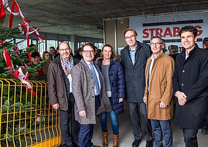 Foto von freundlich schauenden Personen auf einer Baustelle die neben einem Tannenbaum stehen.  