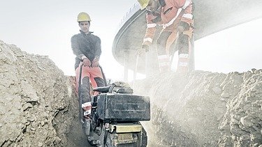 Foto von zwei Bauarbeitern mit einer Maschine bei Tiefbauarbeiten in einem Graben