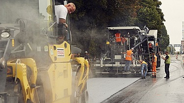 Foto von mehreren Straßenbauarbeitern und zwei Asphaltbaumaschinen bie der Arbeit