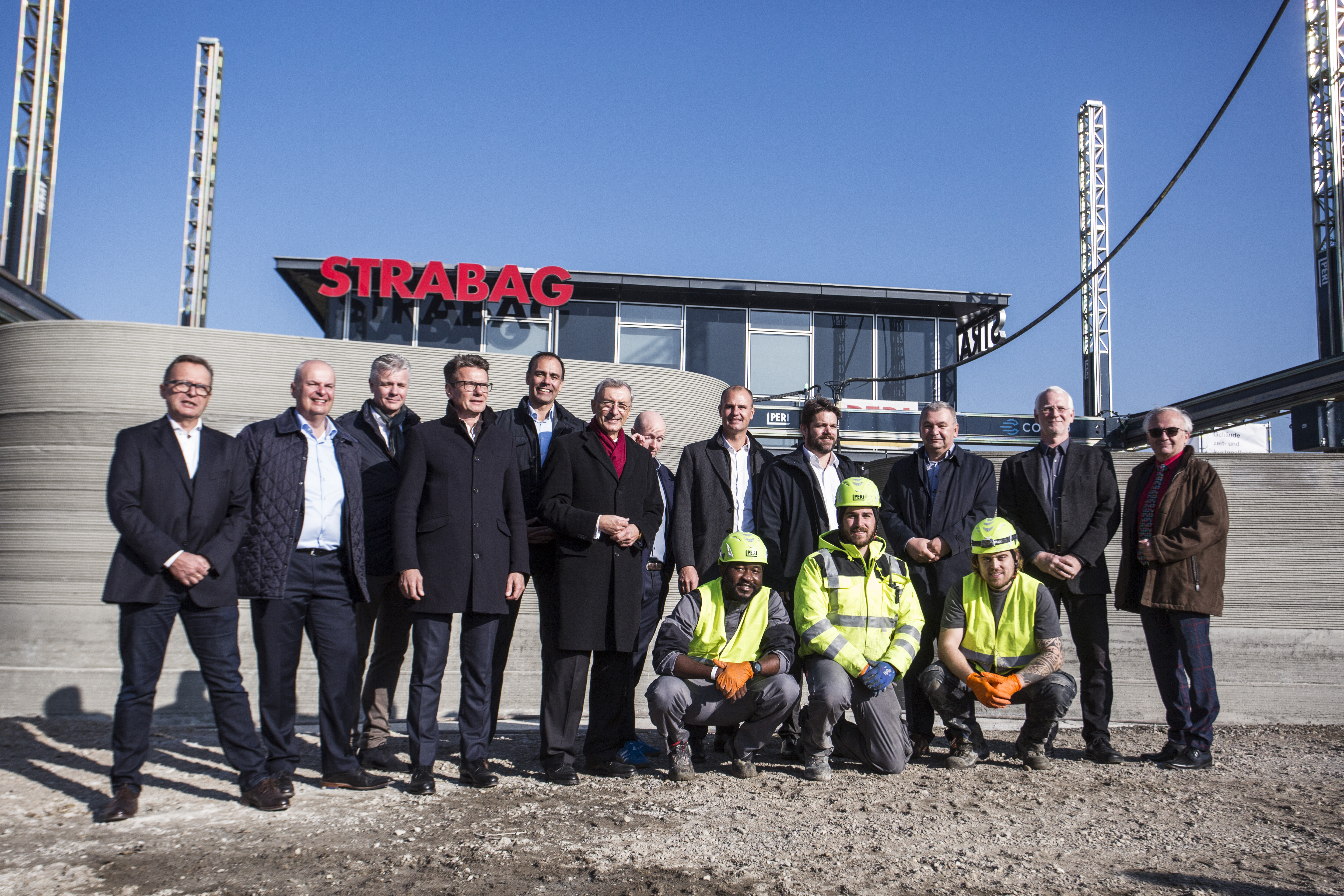 Photo of several people standing in front of a concrete wall, an office building in the background