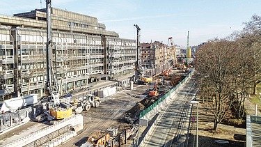 Foto von einer aufgerissenen Straße. Es sind mehrere Kräne und andere Baugeräte zusehen.