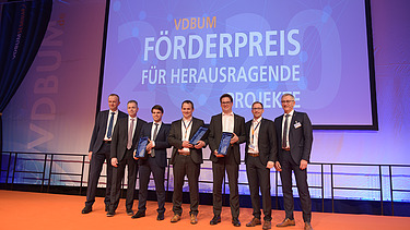Foto von mehreren Männern in Anzug auf einem Bühnen. Im Hintergrund ein Banner vom VDBUM: „Förderpreis für Herausragende Projekte“ zu sehen.            