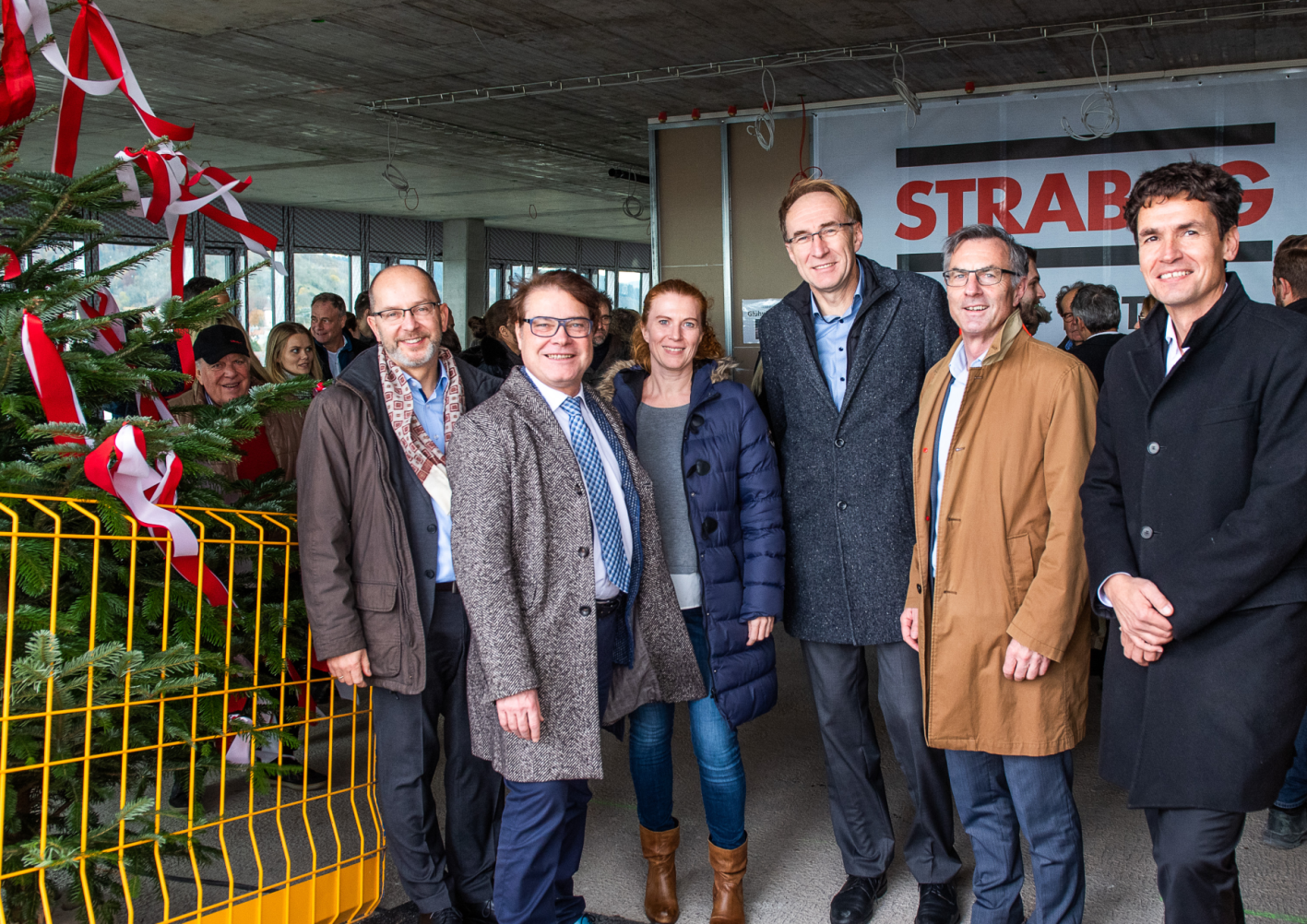 Foto von freundlich schauenden Personen auf einer Baustelle die neben einem Tannenbaum stehen.  