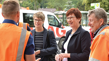 Foto von vier Personen, die linke und die rechte Person sind Arbeiter in Arbeitskleidung, in der Mitte stehen zwei Frauen